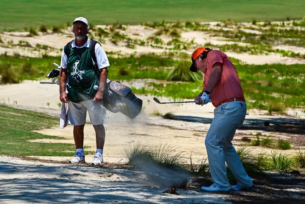 William Holcomb V plays a shot from the native area while caddie Keith Silva looks on.